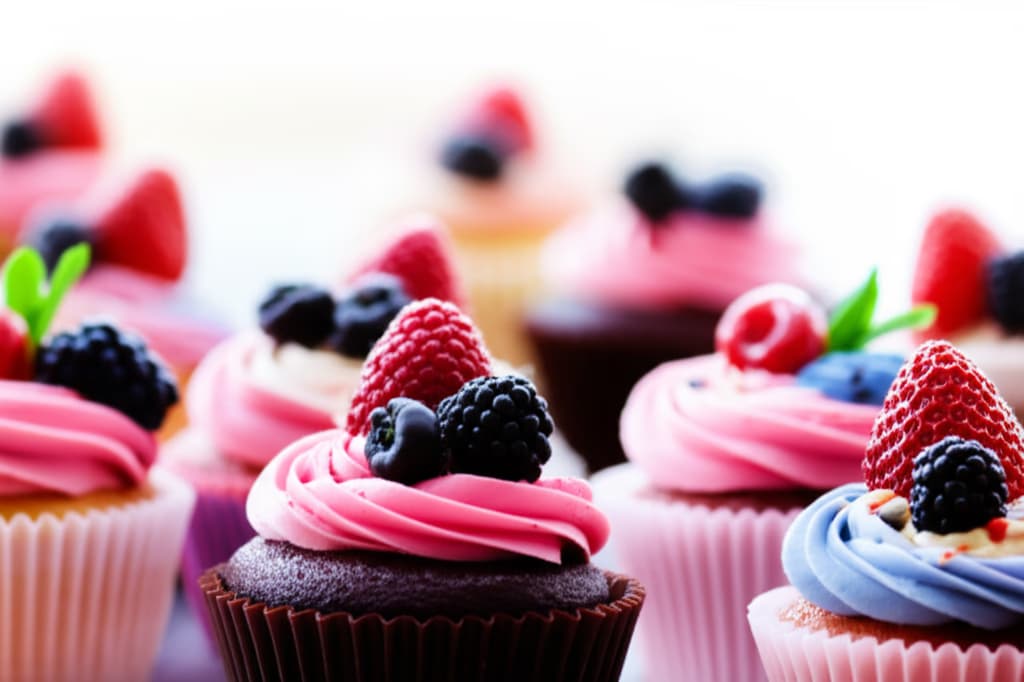 An assortment of beautifully decorated, high-quality gourmet cupcakes with intricate frosting, fresh fruit, and sprinkles, presented on an elegant white stand.